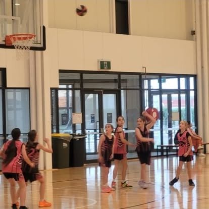 A Panthers junior girls team in pink and black singlets during a game, one player lining up a shot while teammates get into position.